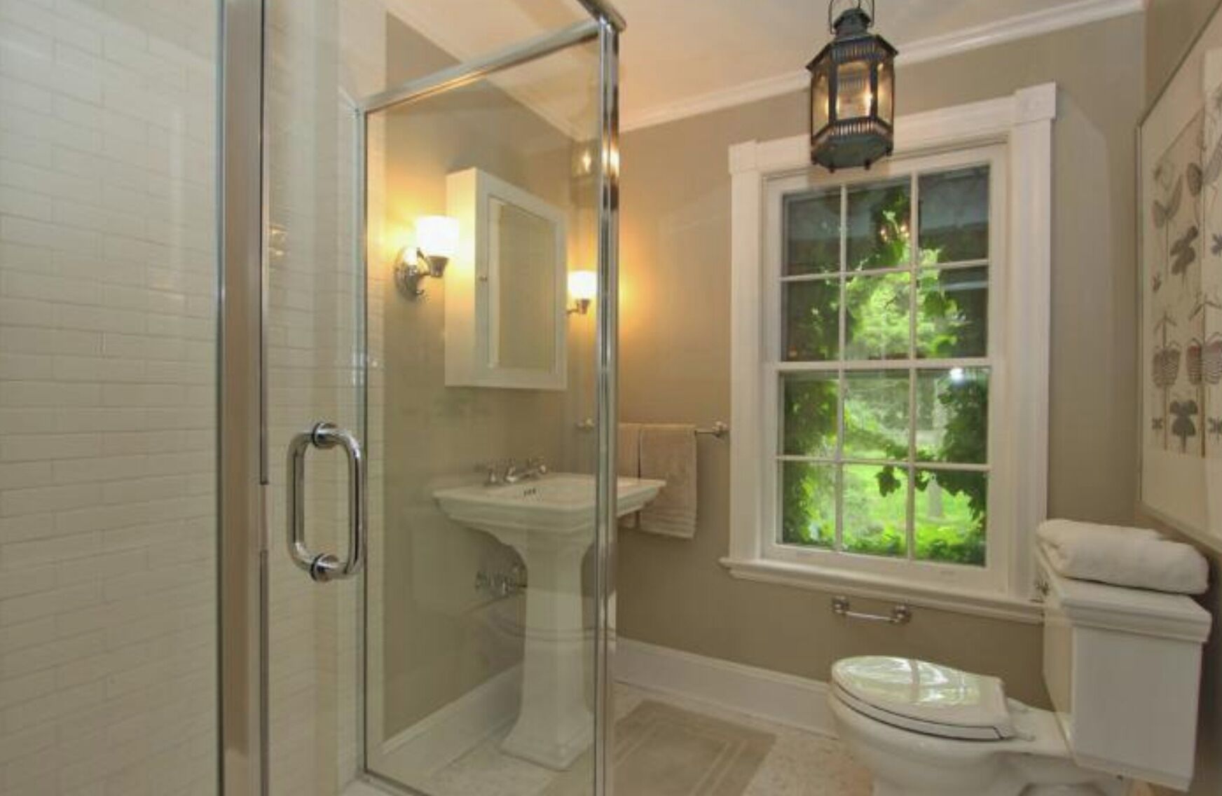 A bathroom with a glass shower enclosure, a sink, a toilet, and a window with a view of greenery.