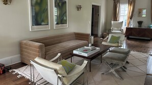 A living room with a beige sofa, white armchairs, a coffee table, and a chandelier. There are two framed pictures on the wall and a door leading to another room. - Spacious Historical 6-bed house in Princeton. Pool, bocce and basketball court. (Princeton)