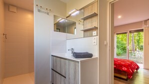 A modern bathroom with a shower, a sink, and a mirror. There is a bed with a red blanket in the adjacent room. - La Salamandre - maison avec jacuzzi (Selles-sur-Cher)