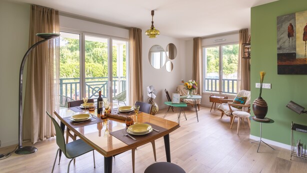 Ein Essbereich mit einem Holztisch, gedeckt für vier Personen, einer Stehlampe und Blick ins Freie durch große Fenster.