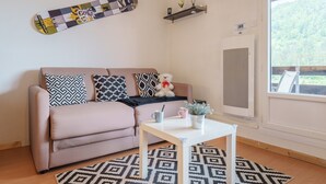 Een woonkamer met een beige bank, gekleurde kussens, een witte salontafel, een skateboard aan de muur en een raam met uitzicht op groen.