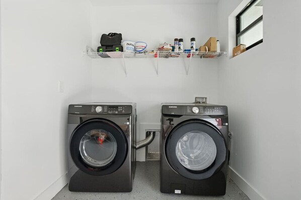 A laundry area with two Samsung washing machines, a shelf with cleaning supplies, and a window. - Brand New Vacation Home! 3/2 (Lehigh Acres)
