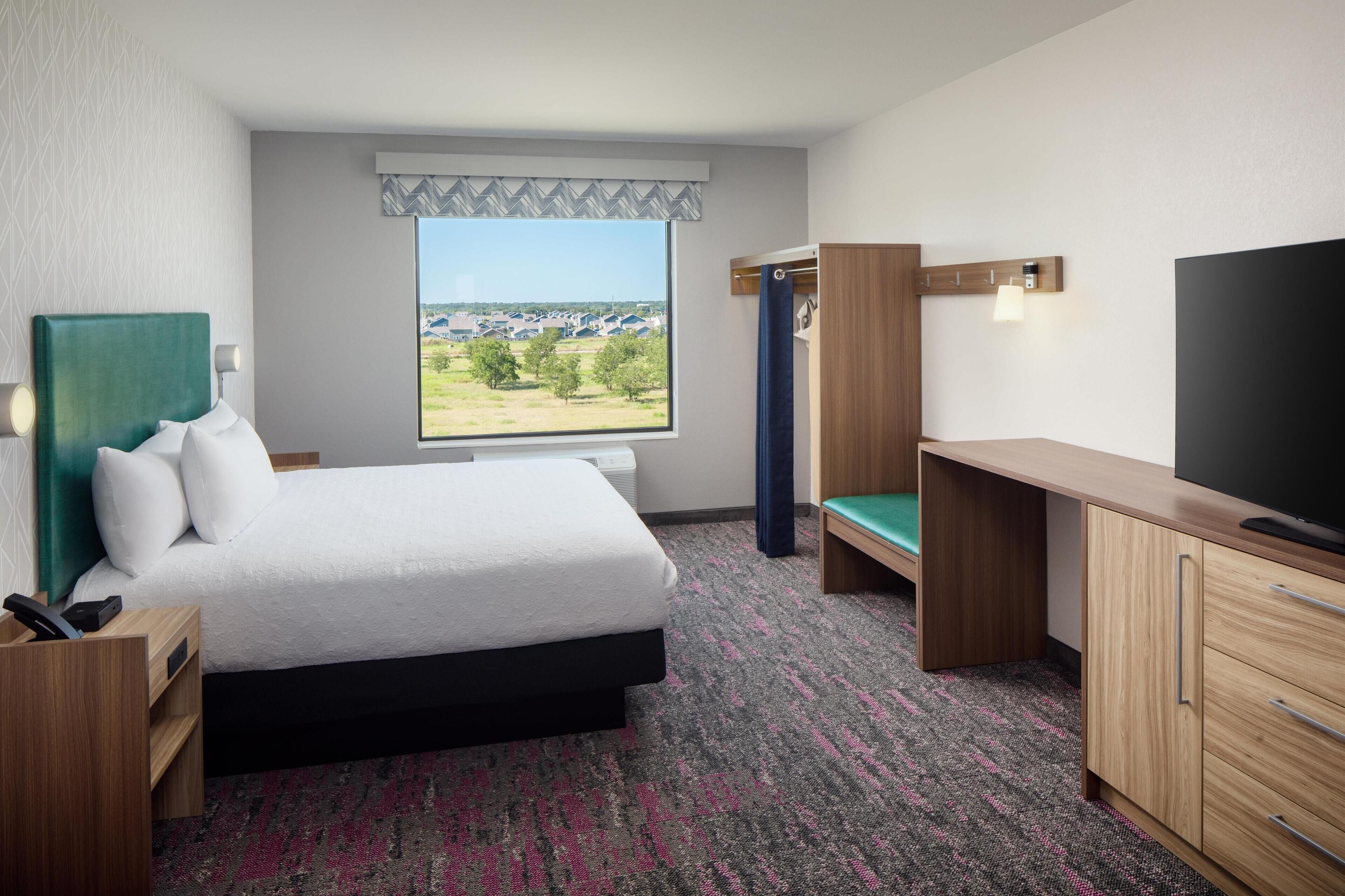 Habitación de hotel con cama, escritorio, televisión y ventana con vistas a edificios.