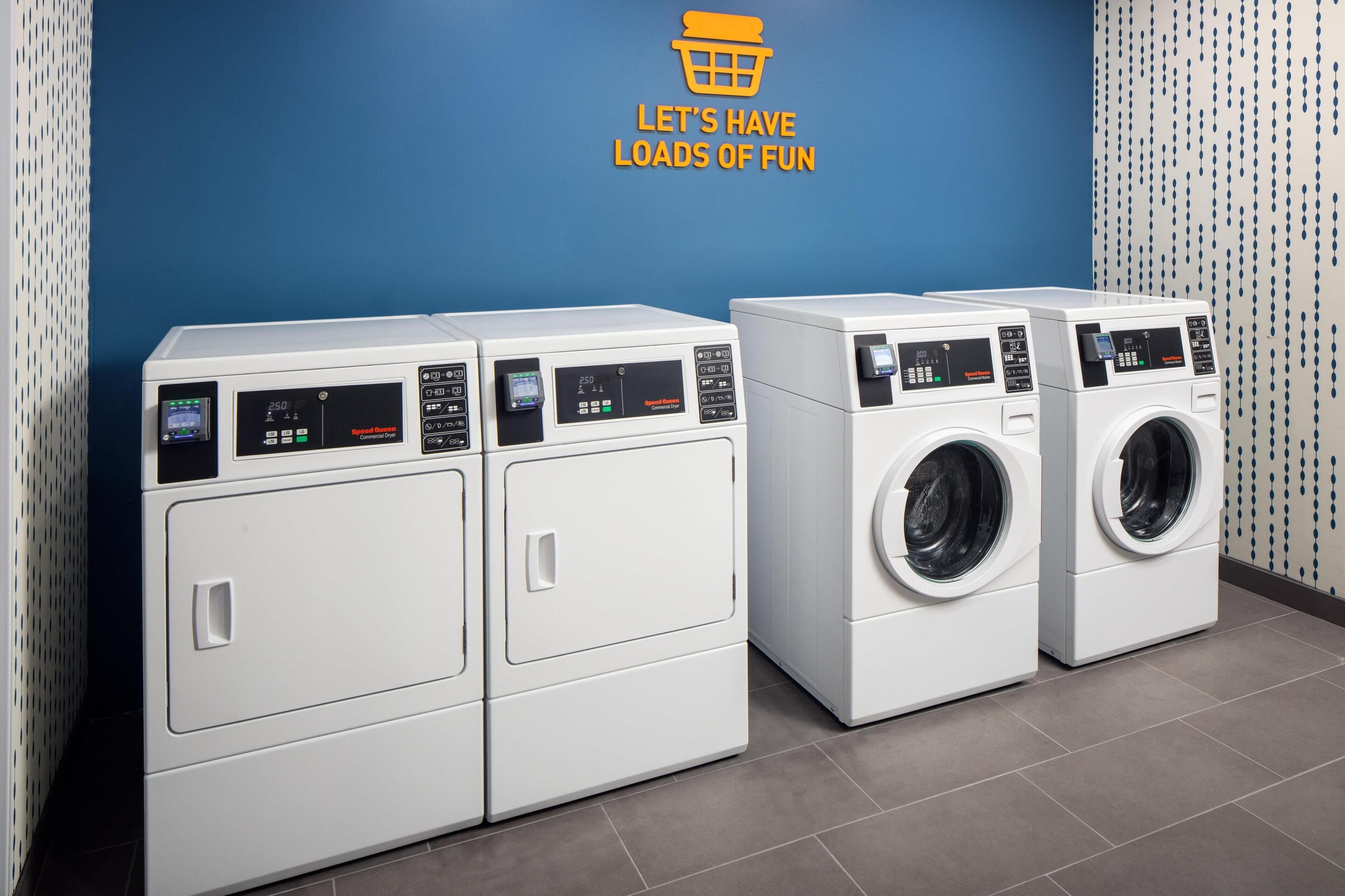 Une buanderie avec trois machines à laver et trois sèche-linge superposés, un mur bleu et un panneau sur lequel on peut lire : « AMUSONS-NOUS ! »