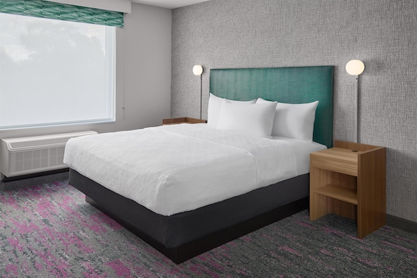 A hotel room with a large bed, a nightstand, a wall-mounted lamp, and a window with blinds. - Home2 Suites By Hilton Frankenmuth (Frankenmuth)