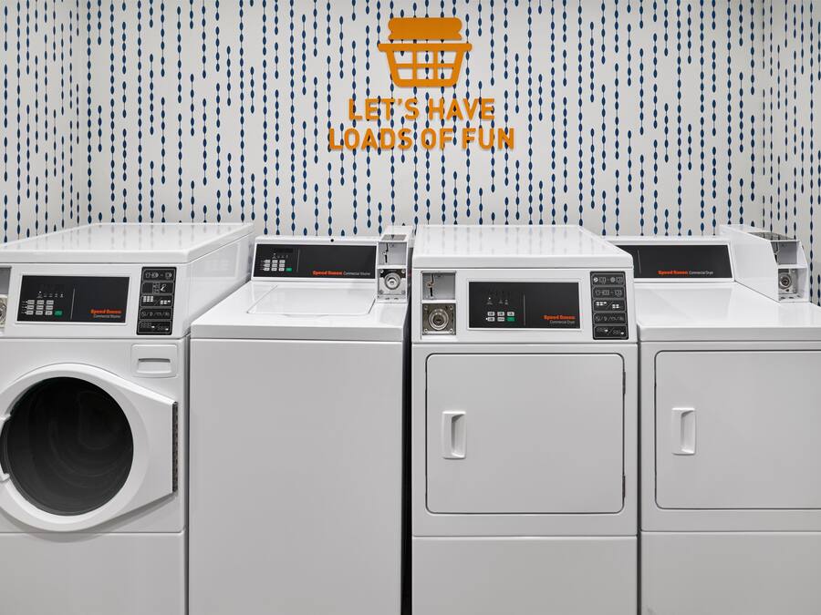 A row of white commercial washers and dryers with control panels, against a wall with a blue and white pattern.