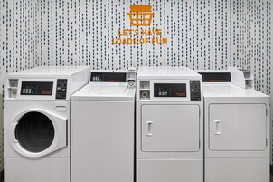 Fila de lavadoras y secadoras comerciales blancas con paneles de control, frente a una pared con diseño azul y blanco.