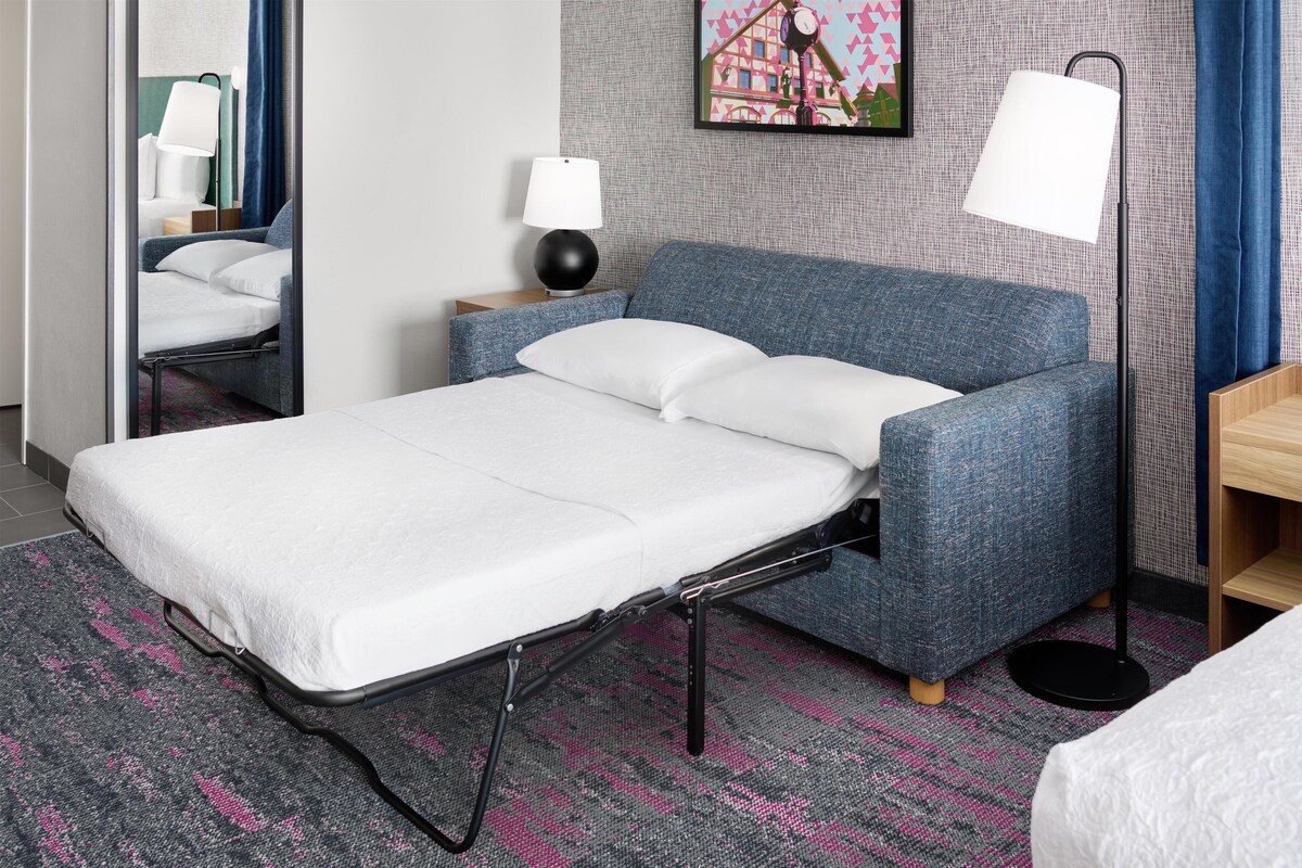 Habitación de hotel ordenada con una cama individual, una mesita de noche con lámpara, un cuadro colgado en la pared y una alfombra estampada.