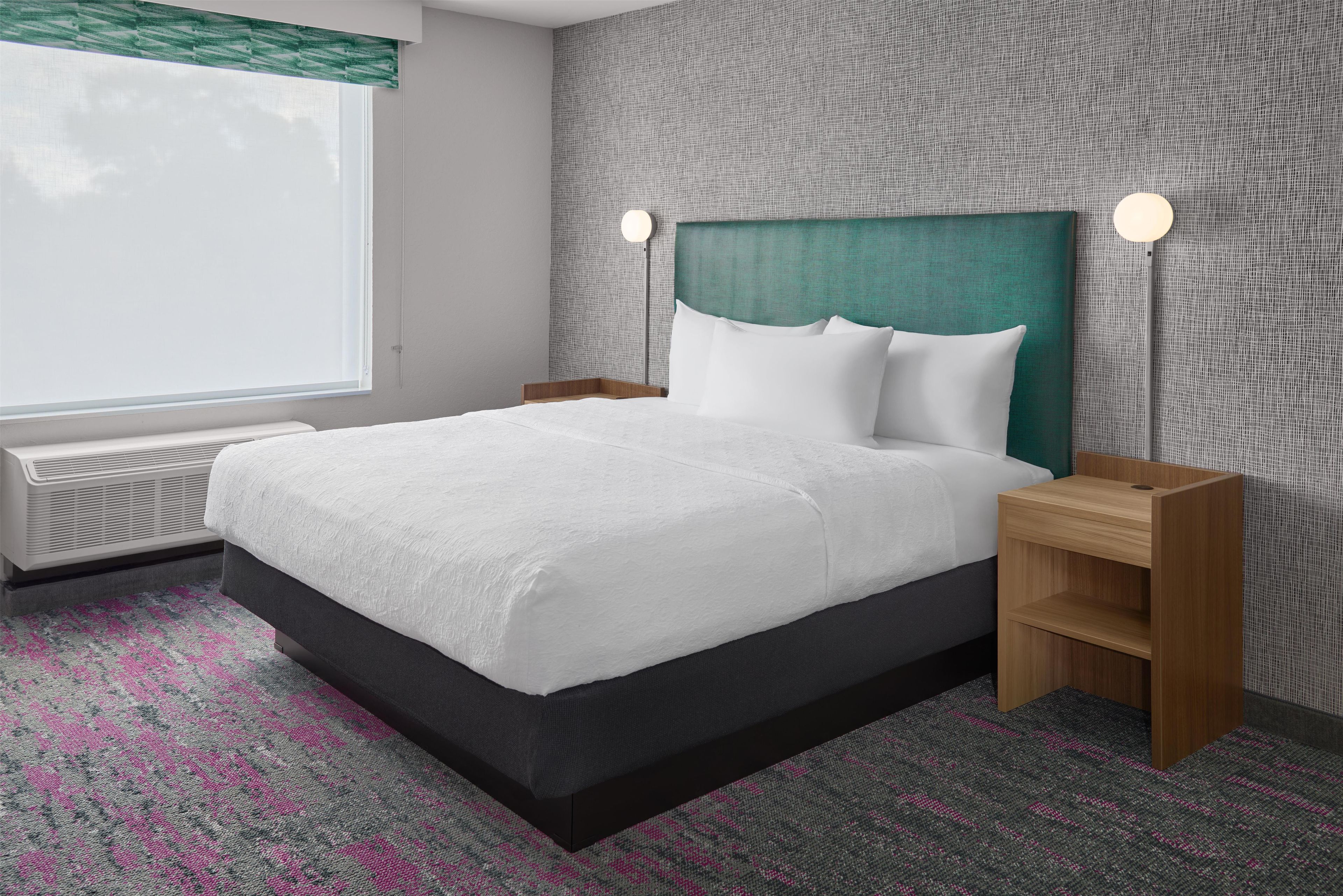A hotel room with a bed, bedside tables, a window with blinds, and a wall with a textured pattern.