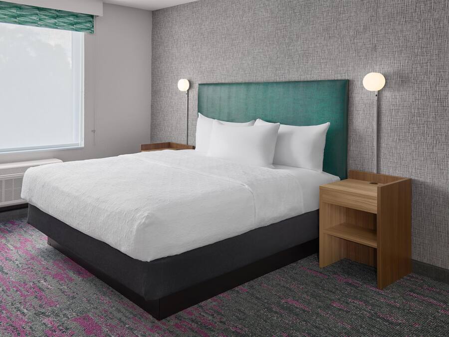 Habitación de hotel con cama, mesitas de noche, ventana con persianas y pared con textura.
