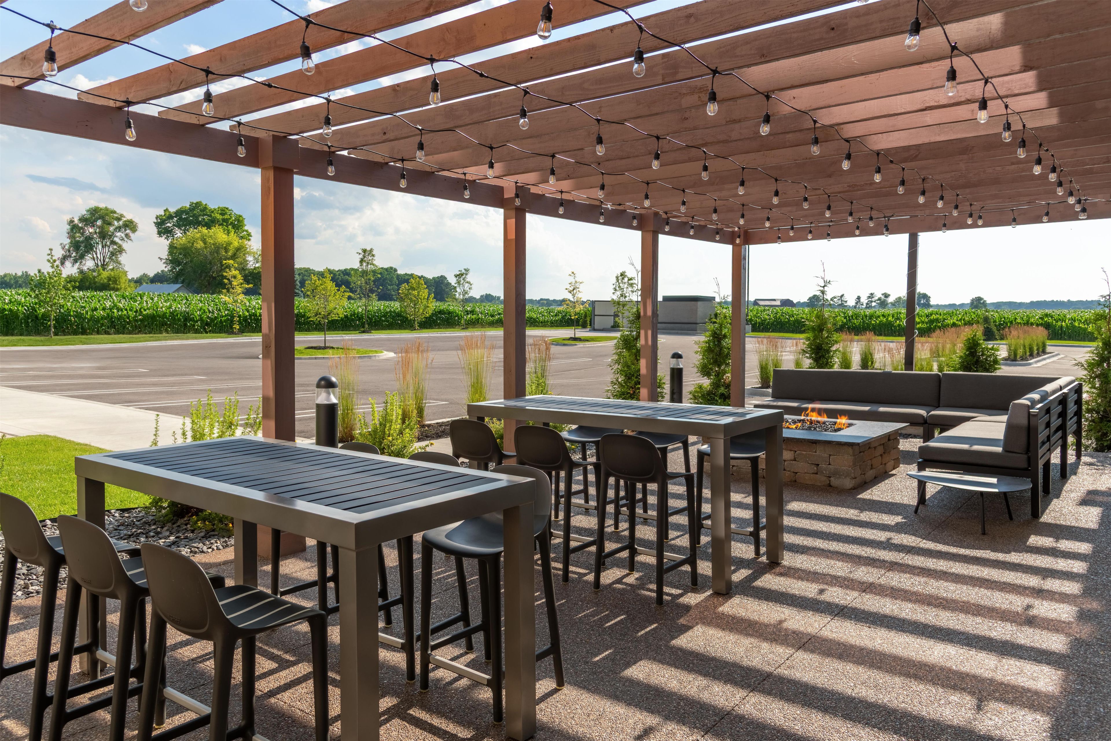 Un patio al aire libre con mesas y sillas, un hogar para fogata y vista a un campo de maíz.