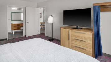 A hotel room with a bed, a TV on a wooden cabinet, a desk with a chair, a mirror, and a floor lamp.
