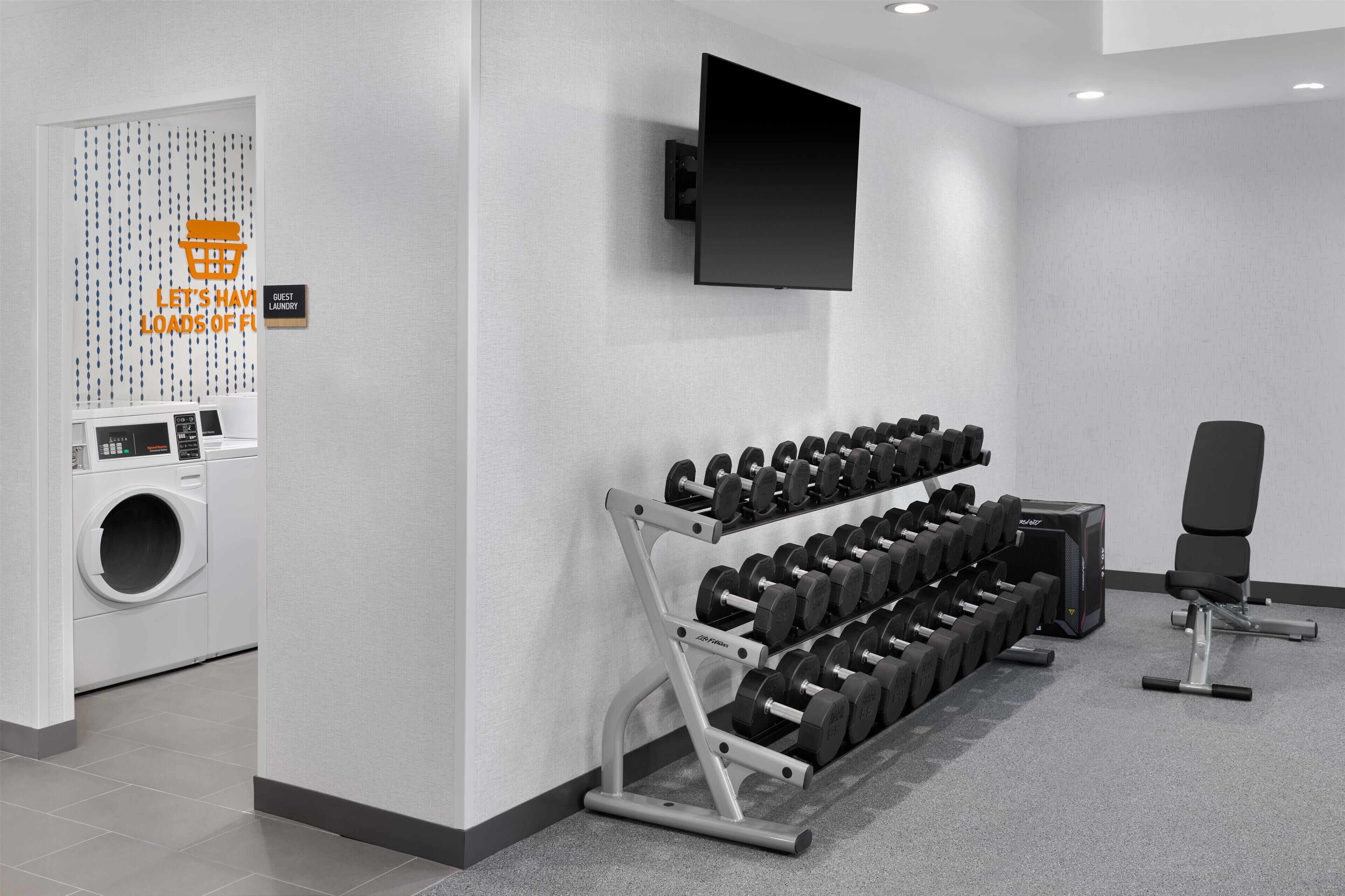 A modern hotel room with a flat-screen TV mounted on the wall, a rack of dumbbells, a black exercise chair, and a washing machine.