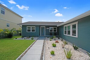 A two-story house with a front porch, a small garden with various plants, and a clear blue sky.