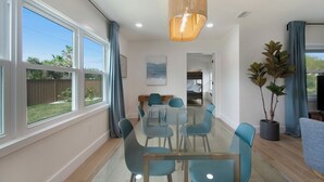 A spacious dining area with a glass table and blue chairs, large windows, and a potted plant.
