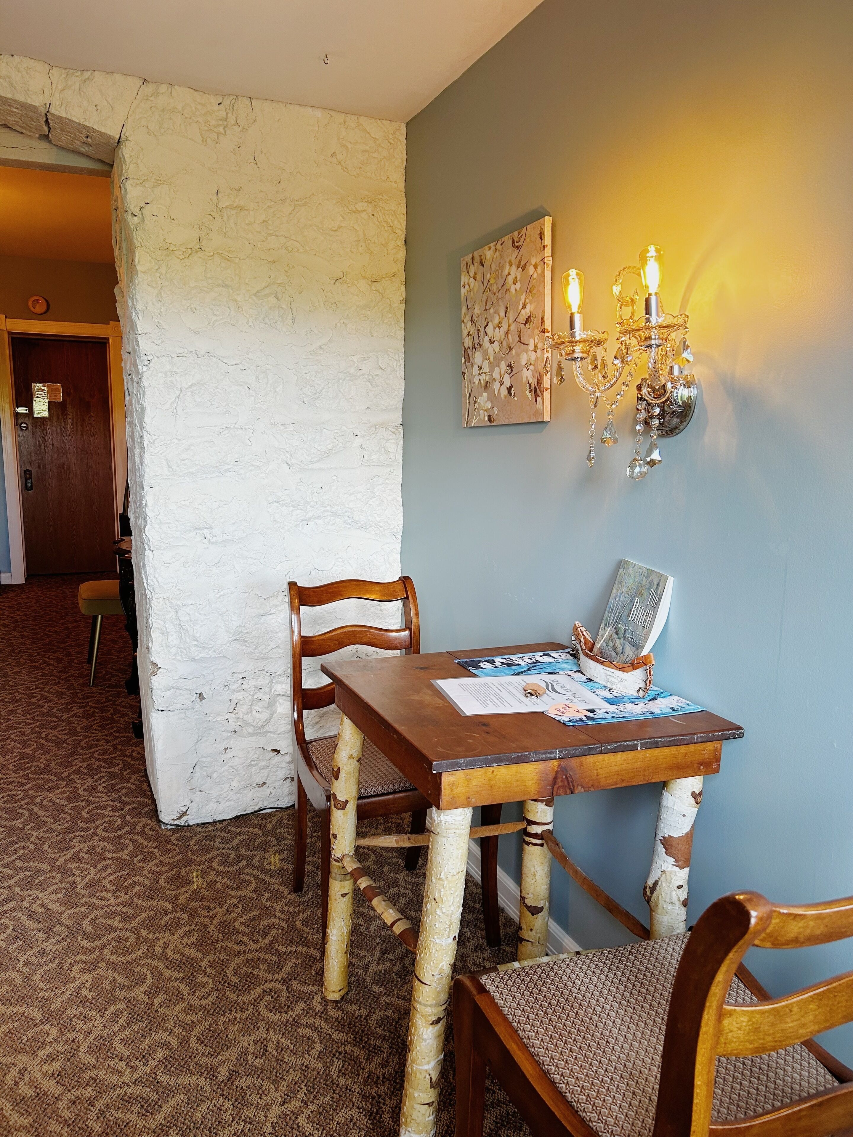 Un comedor con una mesa y sillas de madera rústica, una lámpara fijada a la pared y una puerta al fondo.