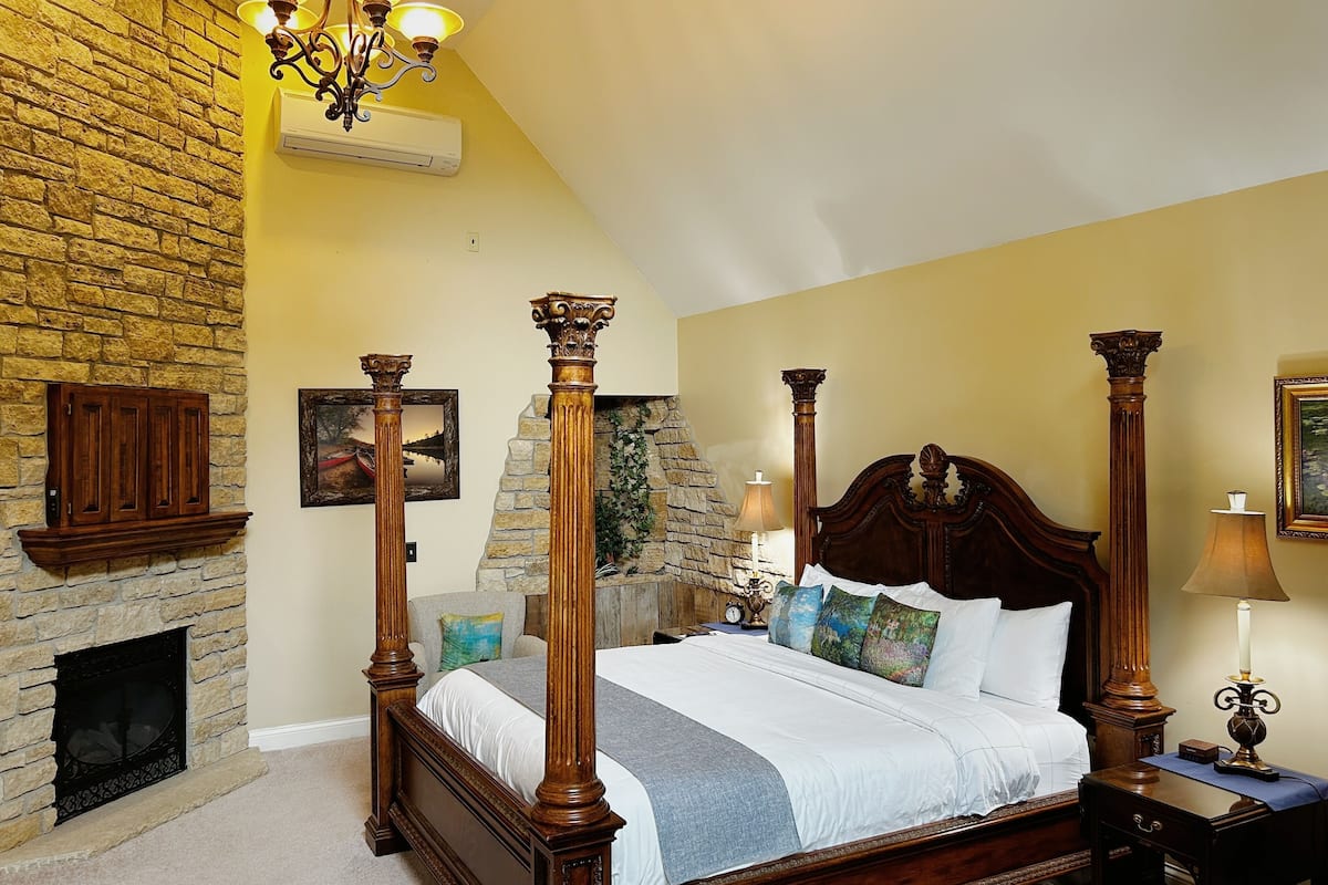 A four-poster bed with a stone fireplace, a chandelier, and a nightstand with a candle.