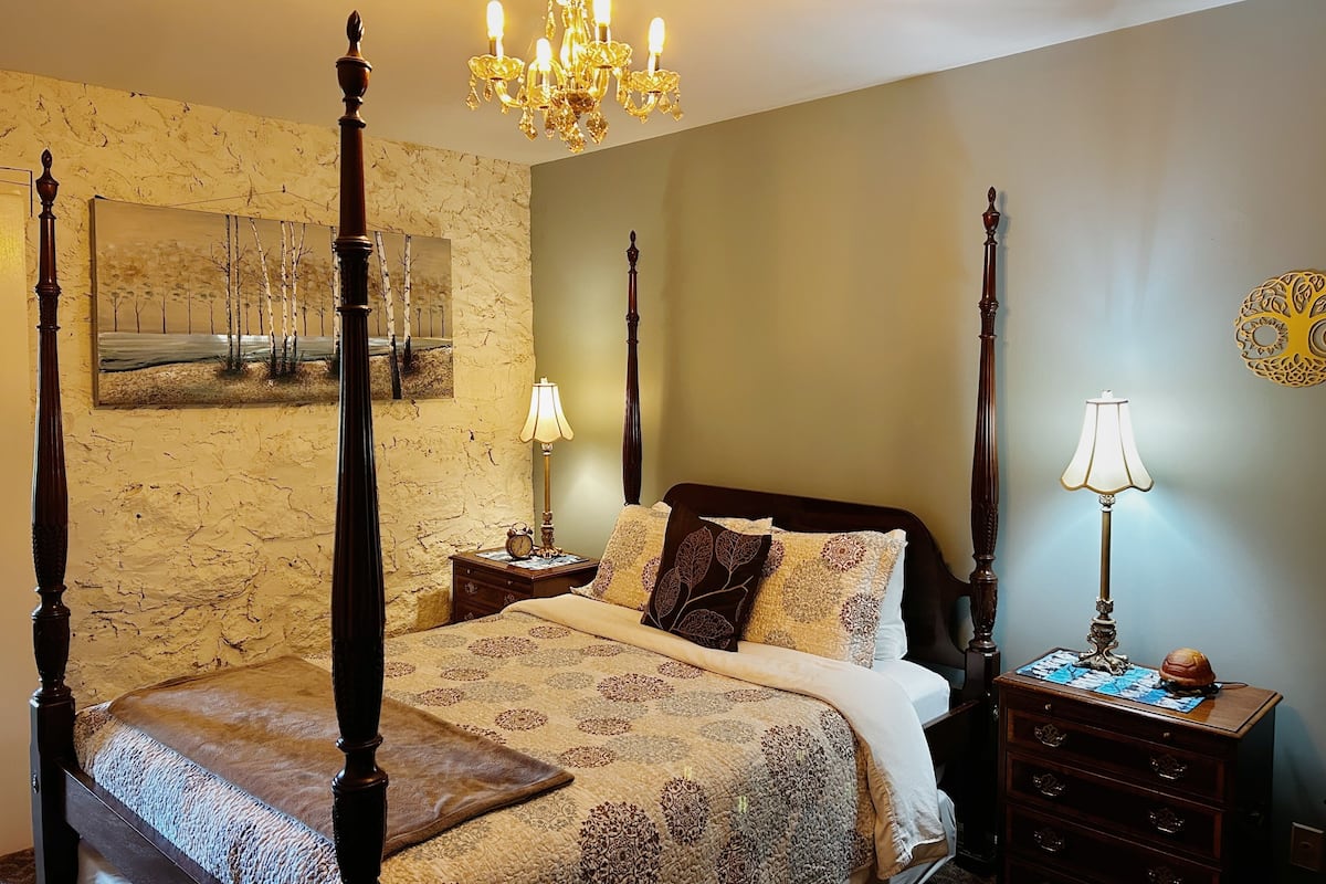 A four-poster bed with a canopy, a chandelier, a bedside table with a lamp, and a wall-mounted clock.