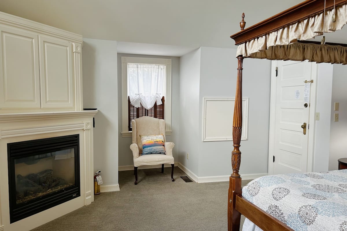 A bedroom with a four-poster bed, a fireplace, a chair with a cushion, and a window with curtains.