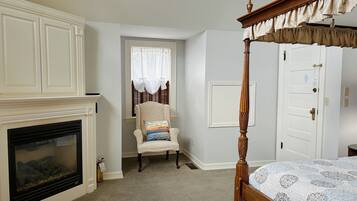 A bedroom with a four-poster bed, a fireplace, a chair with a cushion, and a window with curtains.