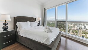 Une chambre moderne avec un grand lit, deux tables de chevet avec des lampes et une vue sur la ville grâce à de grandes fenêtres du sol au plafond.