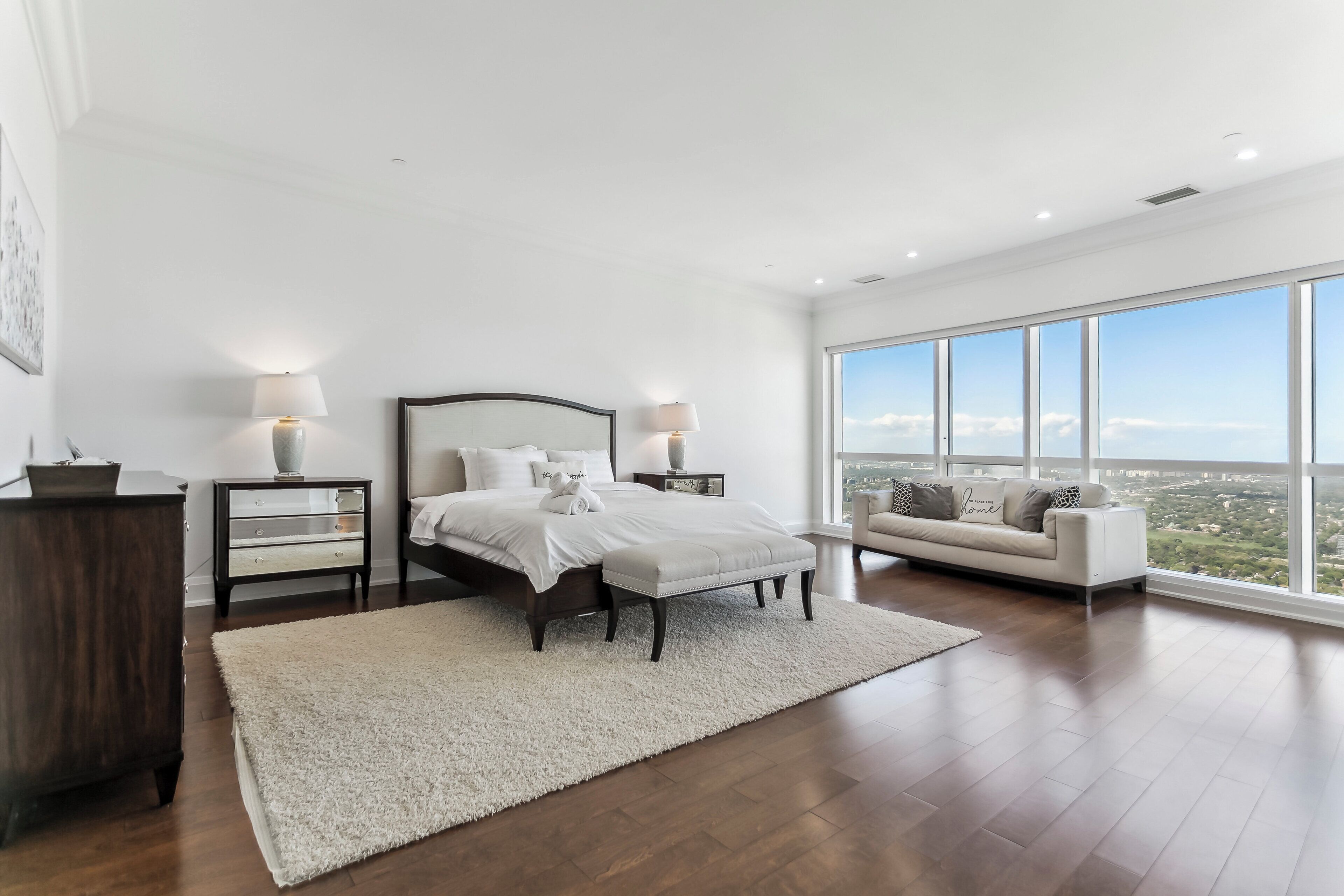 Une chambre spacieuse avec un grand lit, une table de chevet, une commode et une vue sur la ville grâce à de grandes fenêtres.