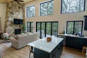A spacious living area with a stone fireplace, a large island, and a view of the outdoors through large windows. - Peaceful Riverside Cabin with Hot Tub. Near WVU & Cheat Lake.  Sleeps 10 (Bruceton Mills)