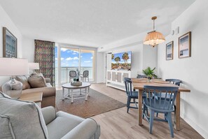 A modern living room with a sofa, a dining table with chairs, a TV, and a balcony with a view.