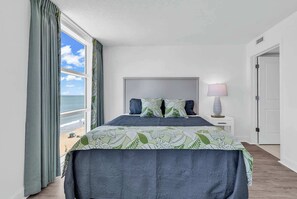 A bedroom with a bed, a nightstand, a lamp, and a view of the beach.