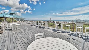 A wooden deck with white tables and chairs, a covered area, and a view of the ocean.