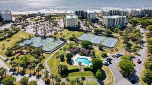 An aerial view of a resort with multiple buildings, a beach, tennis courts, and a swimming pool.