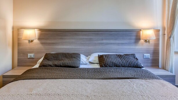 A neatly made bed with a wooden headboard, two bedside lamps, and a window with curtains. - B&B Le Suite di Via Catone (Rome)