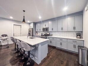 A modern kitchen with grey cabinets, a central island with bar stools, stainless steel appliances, and a hanging light fixture.