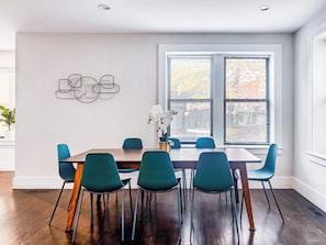 A dining area with a wooden table, teal chairs, a vase with white flowers, and a window with blinds. - Roof Deck, Sleeps 10, W/D, Waterviews! (Boston)