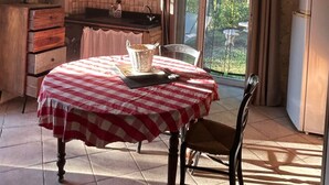 A rustic kitchen with a dining table covered by a red and white checkered tablecloth, wooden cabinets, a hanging light fixture, and a view of greenery outside.