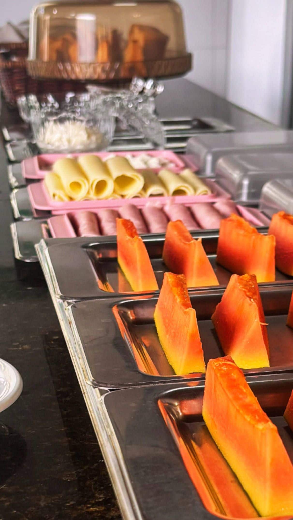 A buffet with various food items including rolled pasta, a glass container with a lid, and orange-colored food items in trays.
