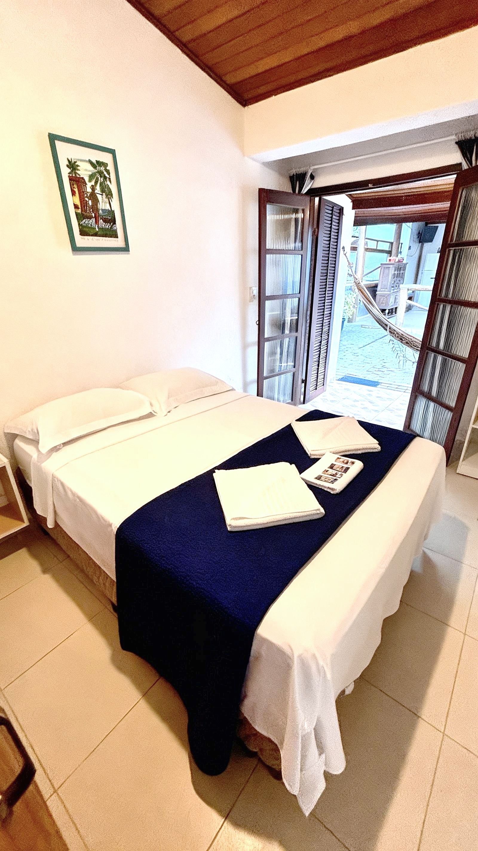 Un lit double avec une couvre-lit bleue et du linge de lit blanc, un tableau encadré au mur et un hamac à l’extérieur de la pièce.
