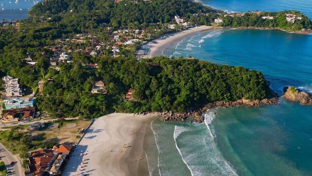 An aerial view of a coastal area with a beach, clear waters, and a forested hill. - Pousada Sr Marinheiro Ubatuba (Ubatuba)