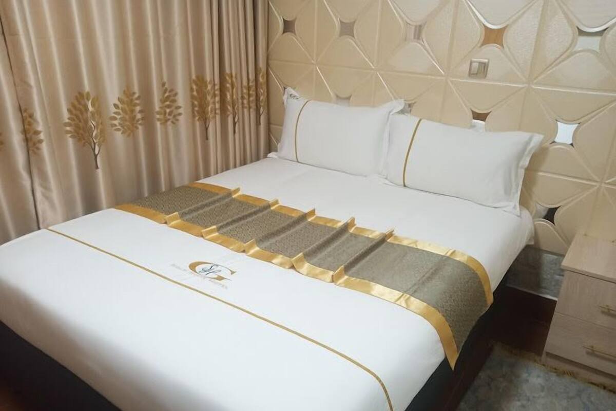 A neatly made bed with a white and gold bedspread, a headboard with a geometric pattern, and a wooden nightstand with drawers.