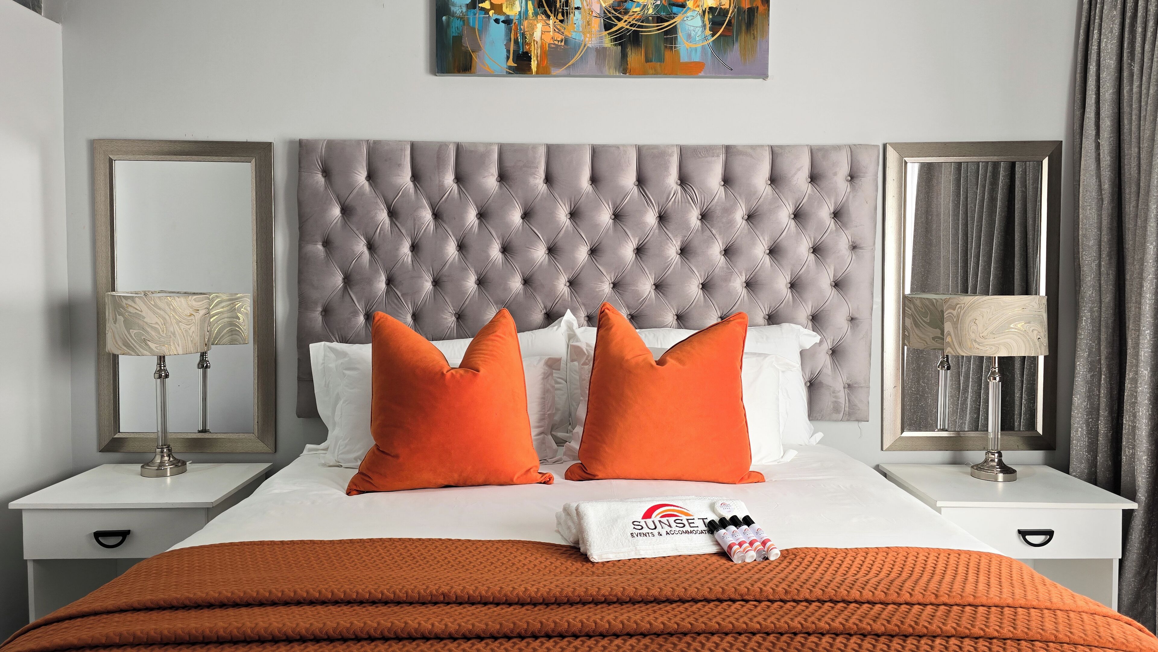 Une chambre d’hôtel avec une tête de lit capitonnée, deux tables de chevet avec des lampes, un lit avec une literie blanche et orange, et un tableau au-dessus de la tête de lit.