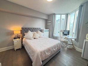 Een slaapkamer met een bed, nachtkastjes, een bureau, een stoel en een raam met uitzicht op de stad.