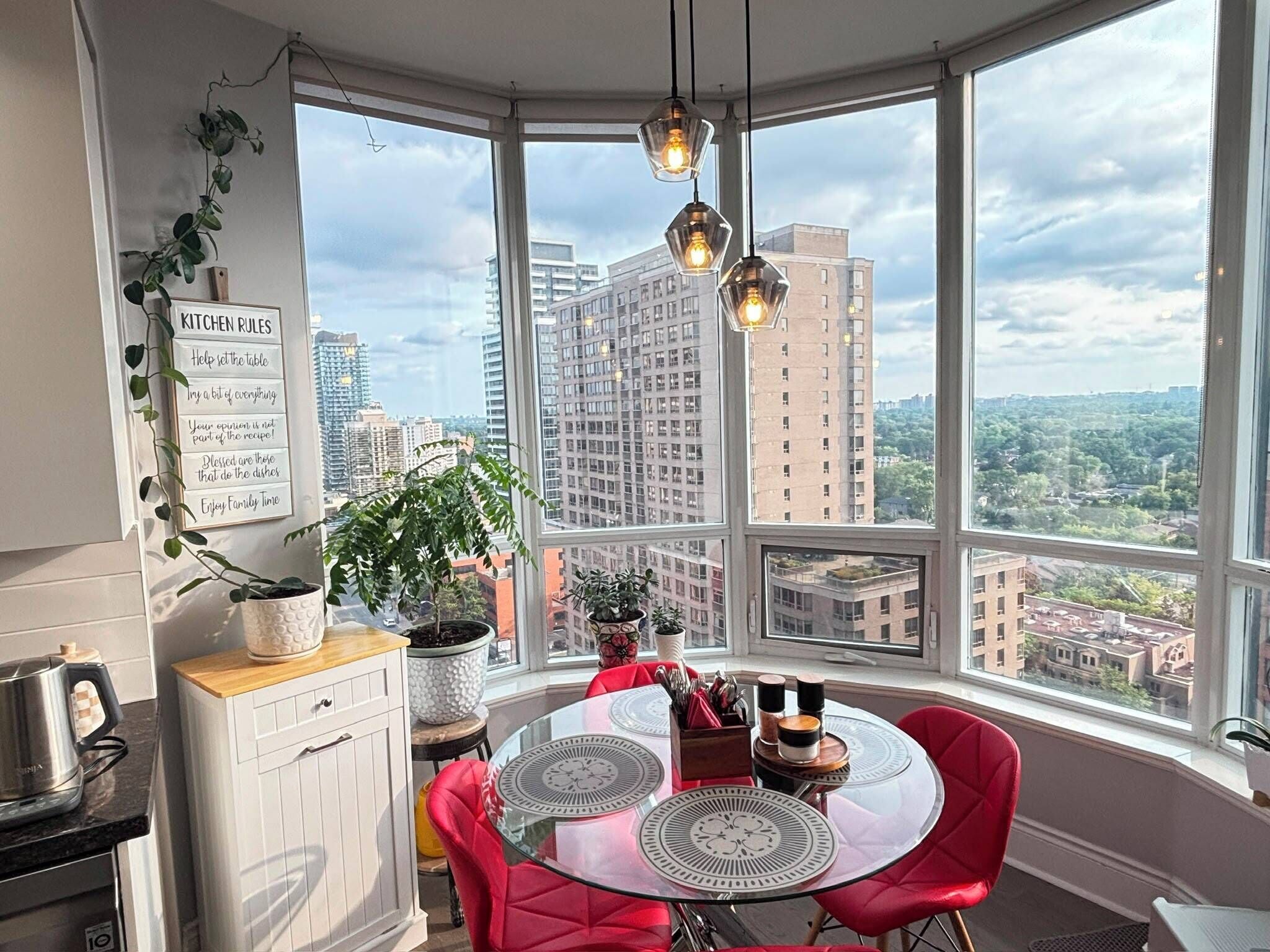 A modern kitchen with a bay window, city view, and a dining area with red chairs.