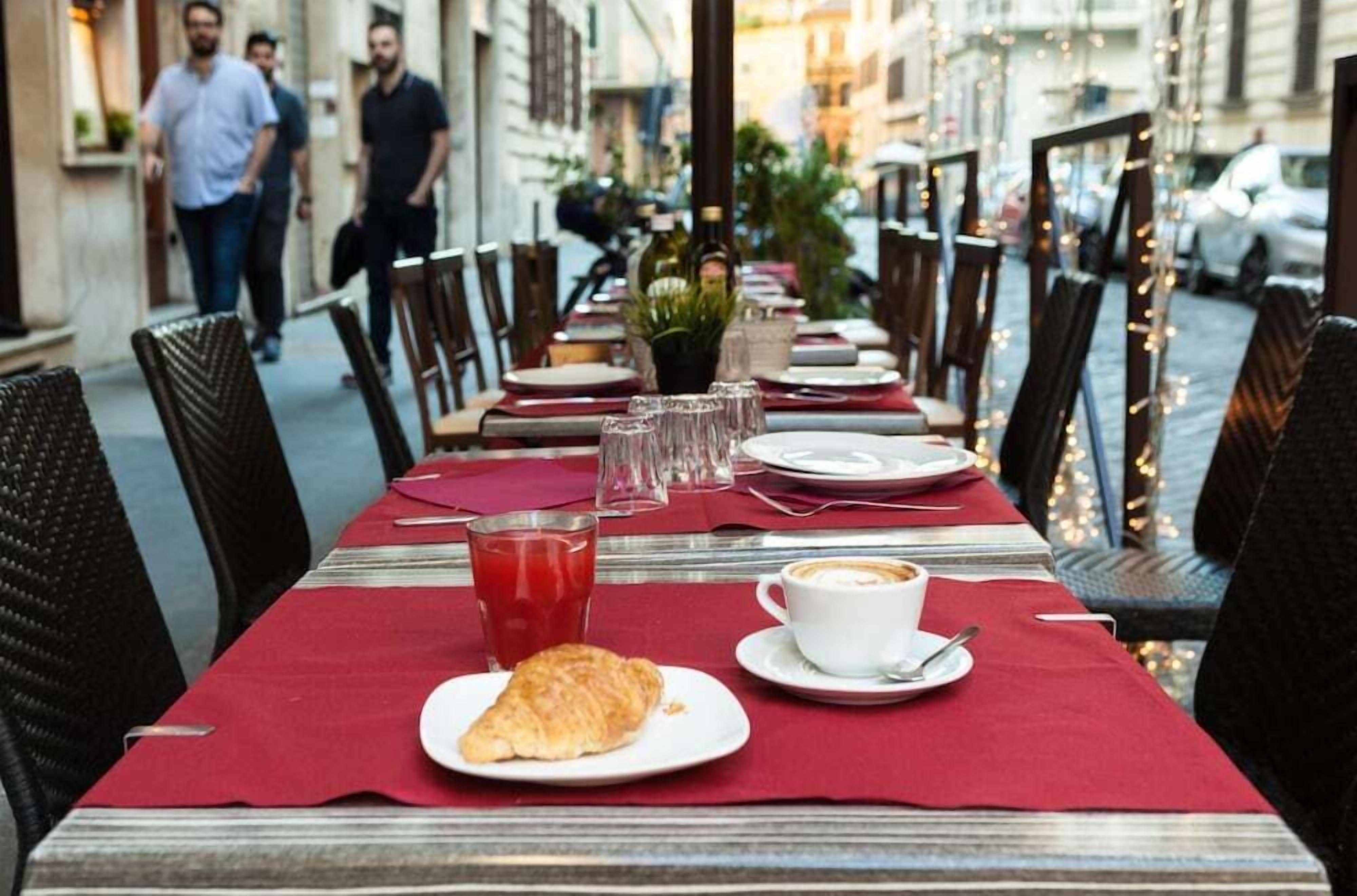 Ruokapöytä on katettu: siellä on croissant, lasi punaista juomaa ja kuppi kahvia. Pöydän ympärillä on tuoleja ja pöydällä on punainen pöytäliina.
