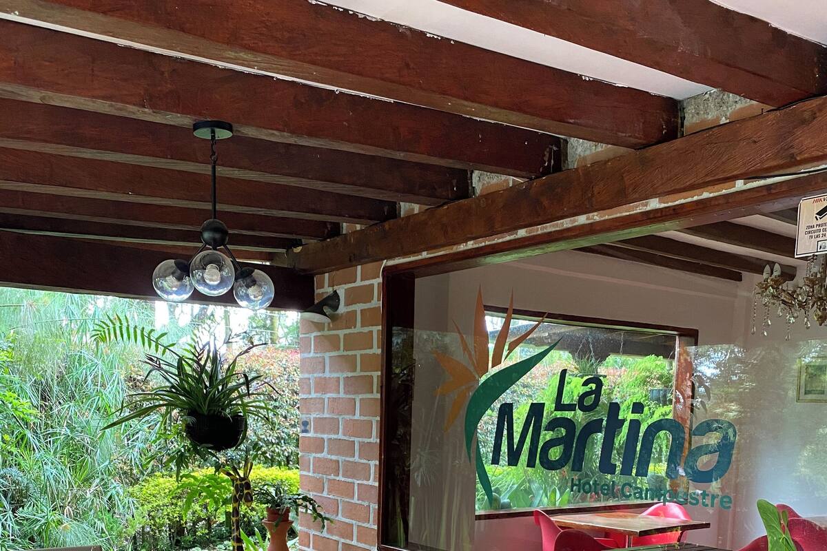 Zona salotto esterna coperta con mobili in vimini, un tavolo in vetro e piante in vaso. Sullo sfondo, un cartello con la scritta 'La Martina Hotel & Country Club'.