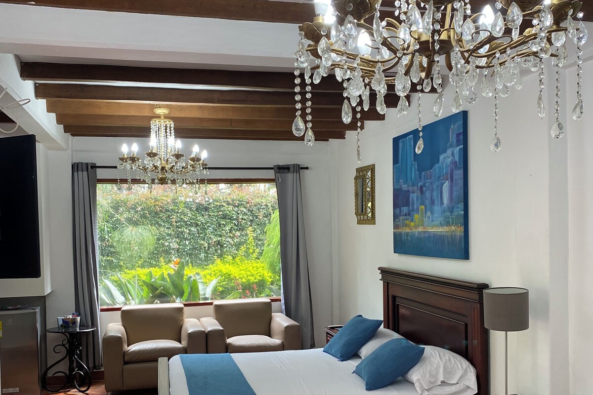 A bedroom with a large bed, a chandelier, a TV, and a view of greenery.