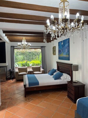 A bedroom with a large bed, a chandelier, a TV, and a view of greenery. - Hotel Campestre La Martina (Rionegro)