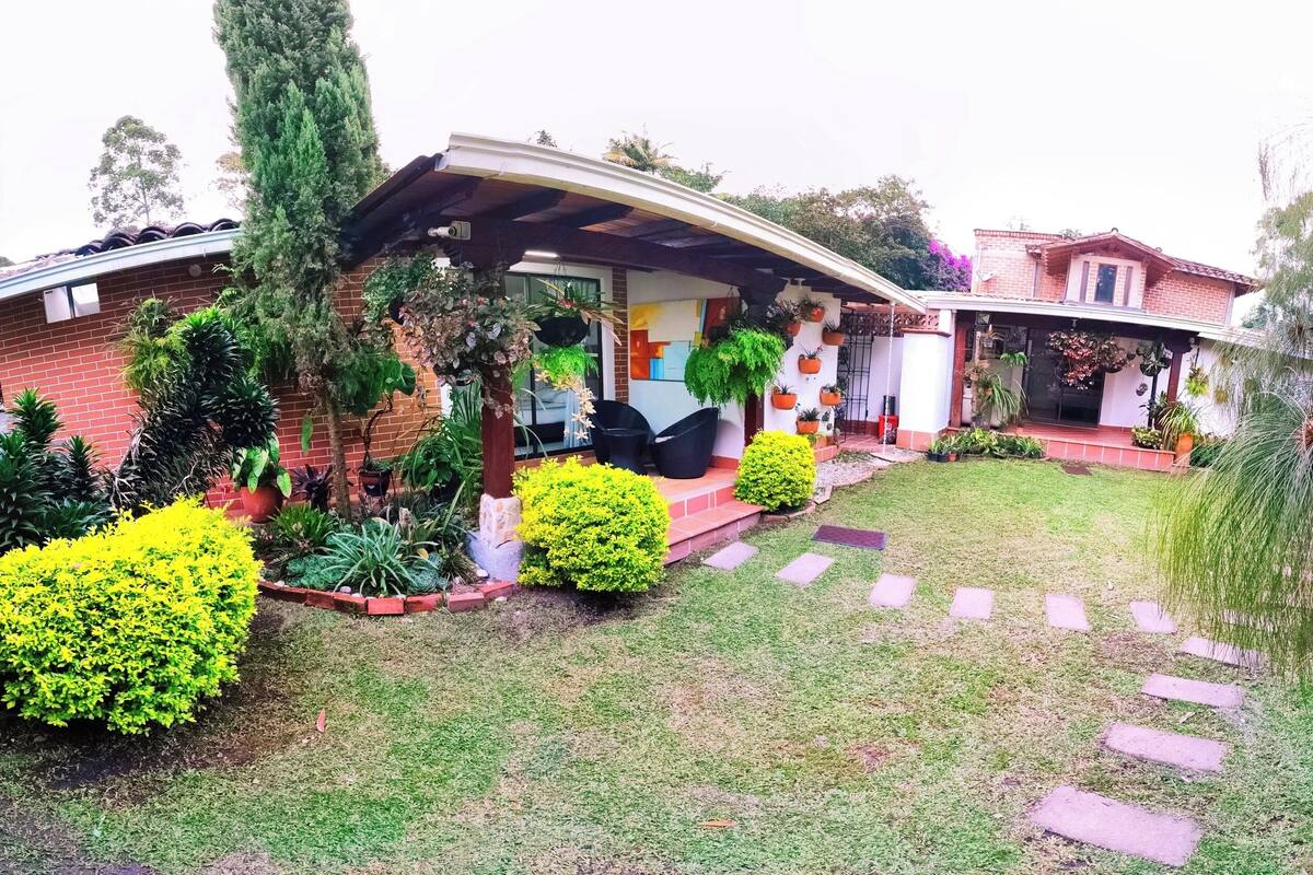 Una casa con tetto curvo, veranda coperta e facciata in mattoni. Nel giardino di fronte si vedono diverse piante e arbusti, e un vialetto che conduce all'ingresso.