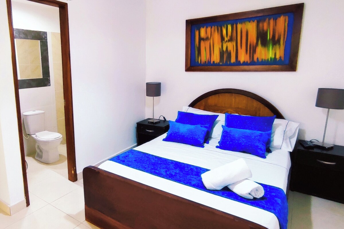 A hotel room with a bed, two bedside tables, a nightstand, a desk, a chair, a wardrobe, and a bathroom.