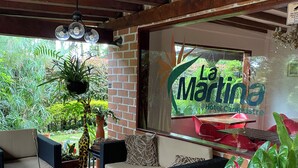 A covered outdoor seating area with wicker furniture, a glass table, and potted plants. A sign for 'La Martina Hotel & Country Club' is visible in the background. - Hotel Campestre La Martina (Rionegro)