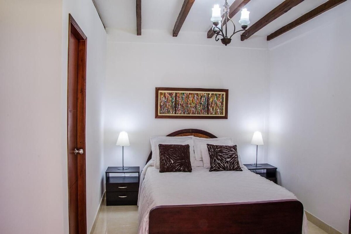 Un dormitorio con cama, dos mesitas de noche con lámparas, techo de madera y un cuadro en la pared.